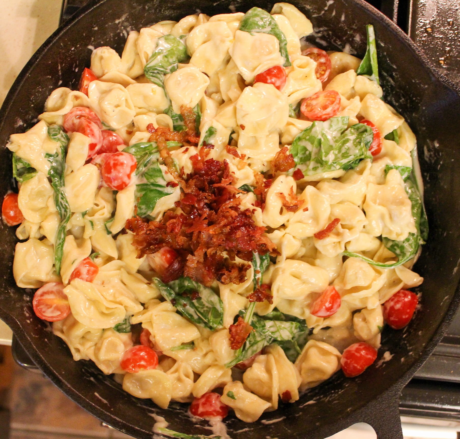 pasta in skillet with cheese, tomatoes, and spinach with cooked chopped bacon on top