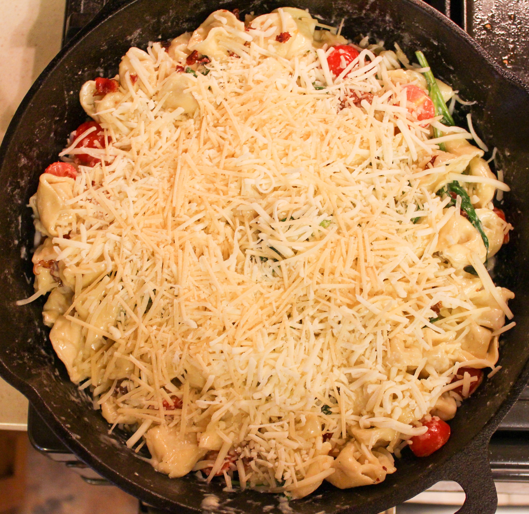 pasta in skillet with cheese, tomatoes, and spinach with cooked chopped bacon on top and cheese on top