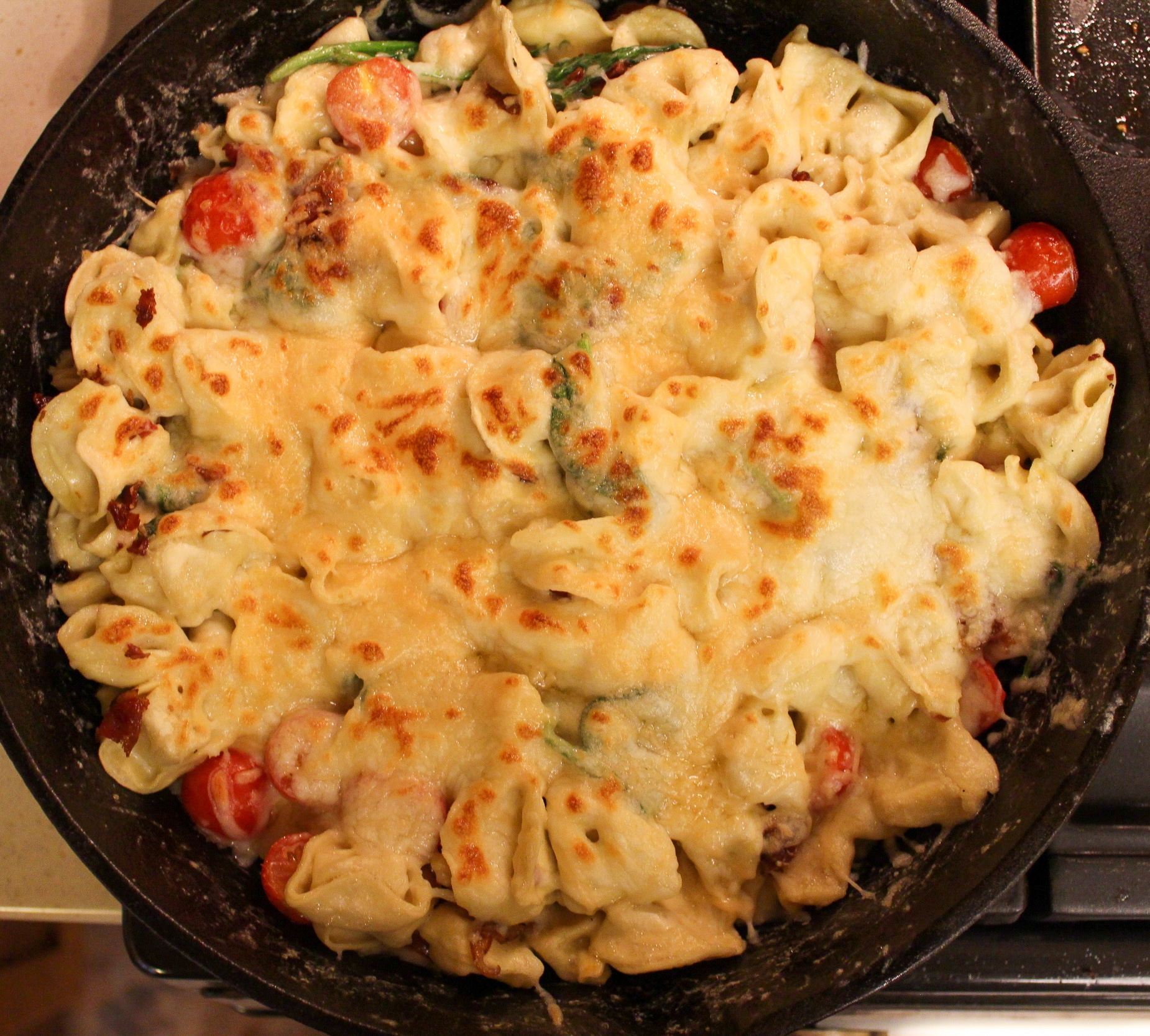 pasta in skillet with cheese, tomatoes, and spinach with cooked chopped bacon on top and cheese on top broiled