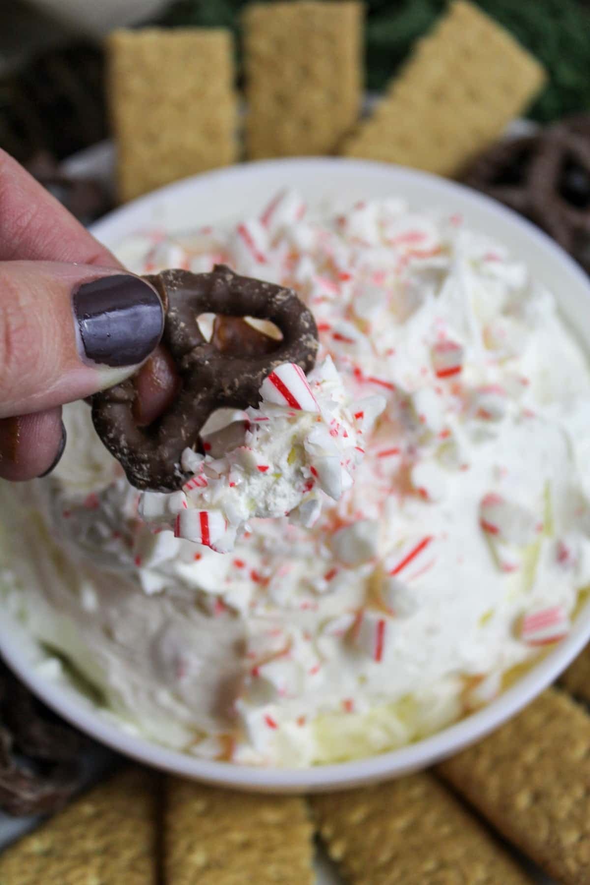 creamy white dip in a white bowl topped with crushed candy canes and surrounded by chocolate covered pretzels. A chocolate pretzel is dipped into the dip.
