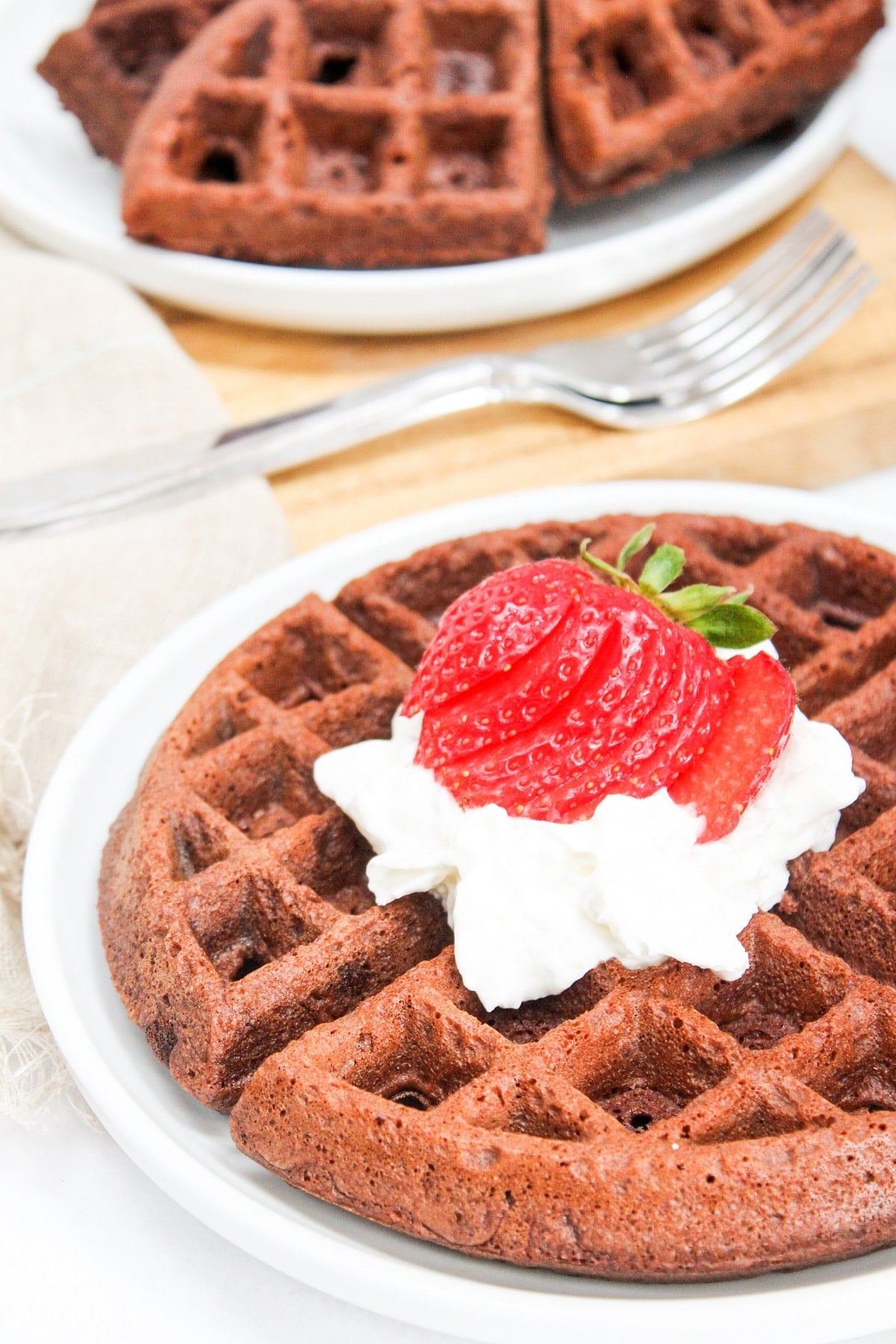 Brownie mix waffles with whipped cream and strawberries on top on a white plate with a plate of waffles in the background
