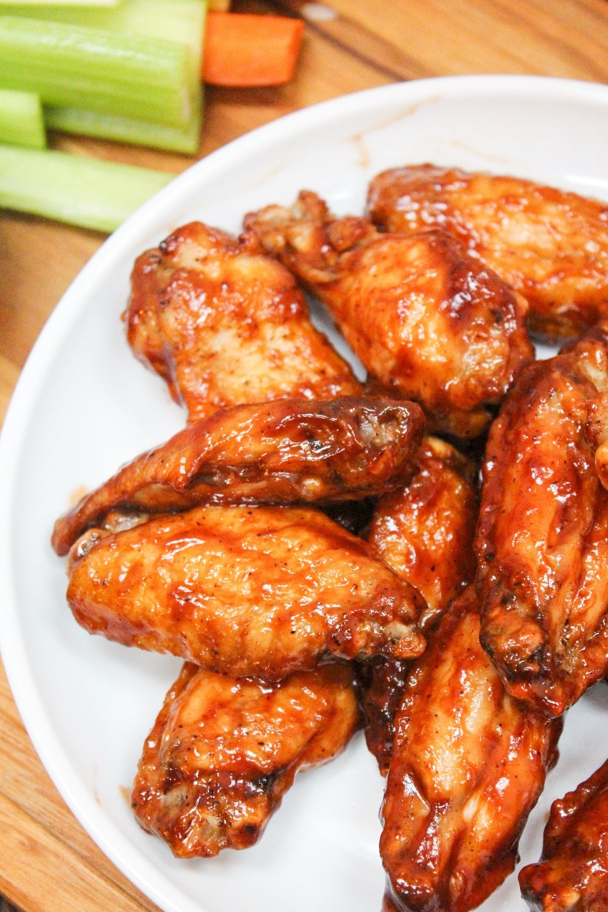 cooked wings in bbq sauce piled on a white plate. Sliced carrots and celery on the side with ranch dressing.