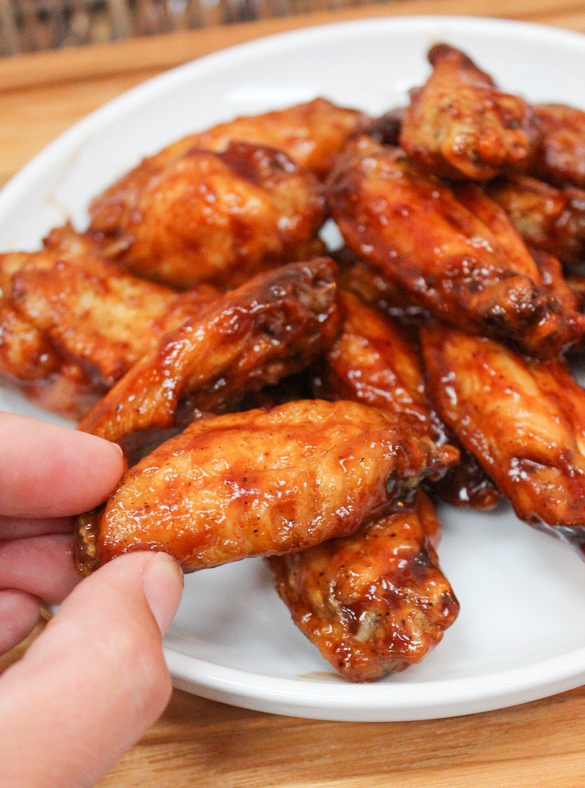 cooked wings in bbq sauce piled on a white plate. Sliced carrots and celery on the side with ranch dressing. One wing is held in hand.
