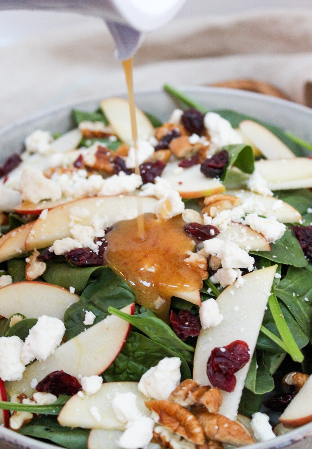 spinach in a gray bowl topped with sliced apples, pecans, crumbled feta cheese, and dried cranberries. Cranberry dressing poured over the top.