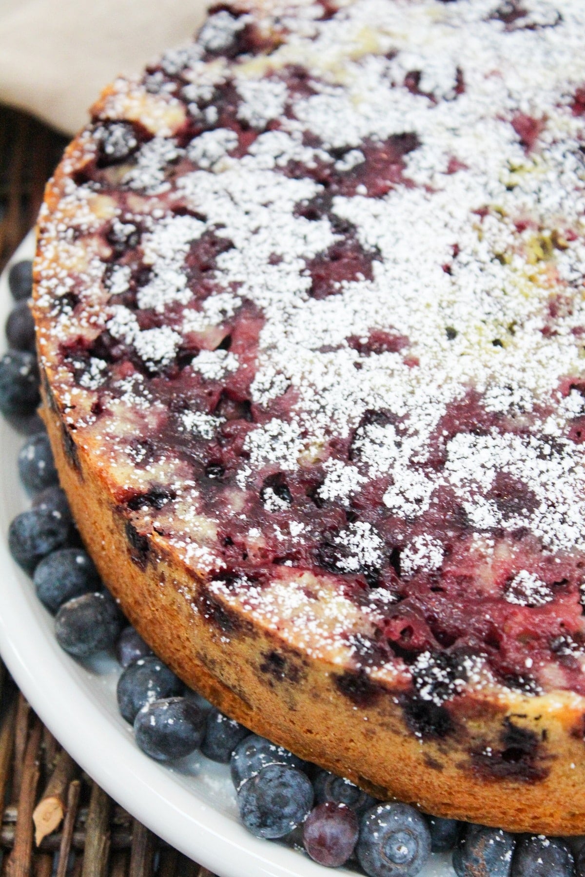 blueberry lemon cake on a white plate topped with powdered sugar and surrounded by fresh blueberries.
