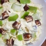Creamy salad in a white bowl topped with chopped snickers bars and fresh green apple pieces.
