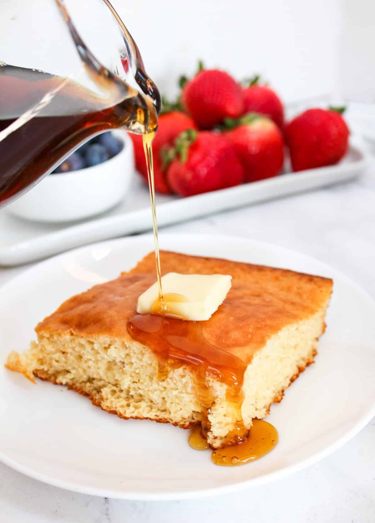 Plain pancake square on a white plate. Topped with butter. Maple syrup is poured from above over the pancake.