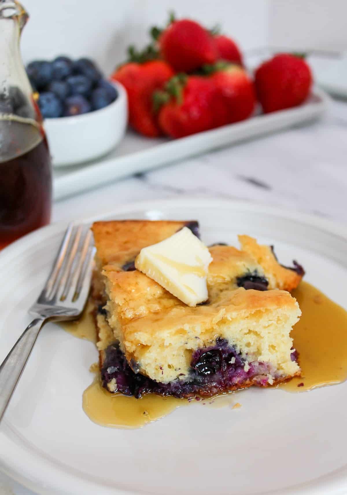 Blueberry pancake square from a sheet pan on a white plate. Topped with butter and maple syrup with a fork on the side.