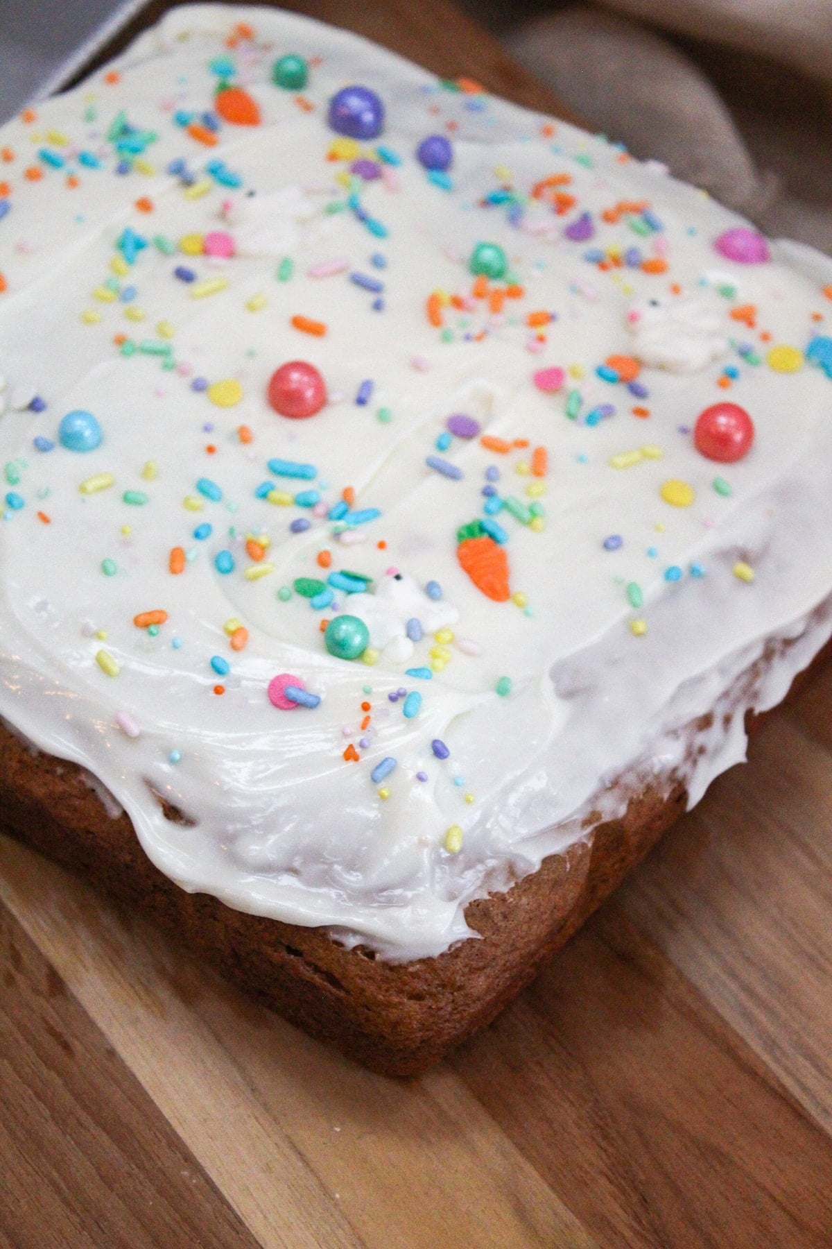 Square cake on a wooden plate. Topped with white frosting and colorful sprinkles.