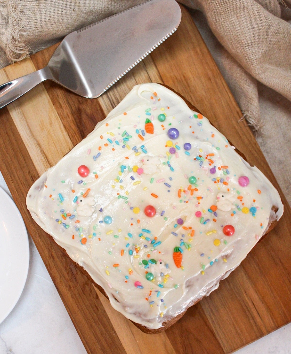 Square cake on a wooden plate. Topped with white frosting and colorful sprinkles.