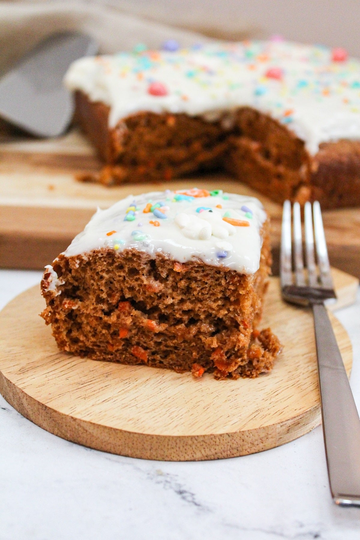 Single slice of carrot cake on a wooden plate with a fork next to it. Topped with white frosting. The larger cake is in the background.